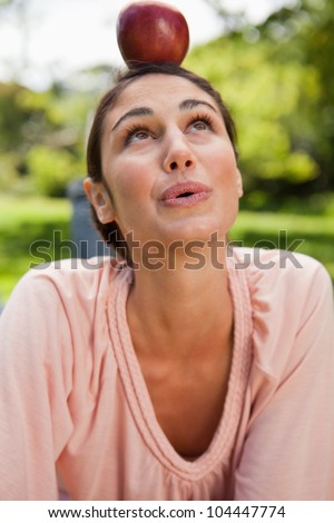 Woman with a concentrated expression balances a red apple on her head while lying prone in grass