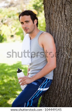 Man with a towel on his shoulder looking towards side while holding a water bottle and resting against the trunk of a tree