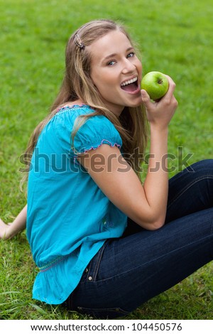Young smiling woman eating a green apple while sitting down on the grass in a park
