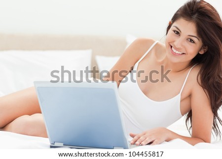 A woman is relaxing on her bed as she types on her laptop while she looks forward and smiles.