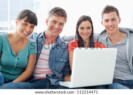 A group of friends sit on the couch together as they hold a laptop and look a the camera