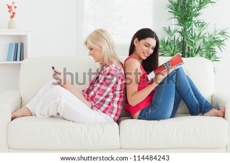 Women sitting back to back on tcouch with magzine and mobile phone