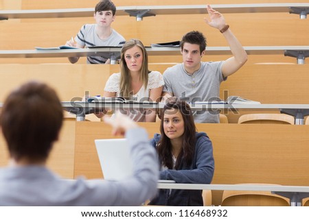 Students sitting at the lecture hall with man raising hand to ask question and lecturer is pointing at him