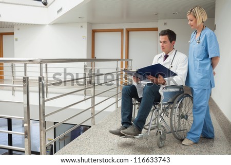 Doctor sitting in wheelchair examining notes with nurse in hospital corridor