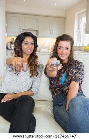 Two women enjoying watching tv holding remote and pointing at screen on sofa