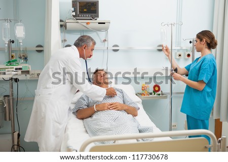 Doctor checking heart beat of patient in bed with stethoscope with nurse adjusting I.V