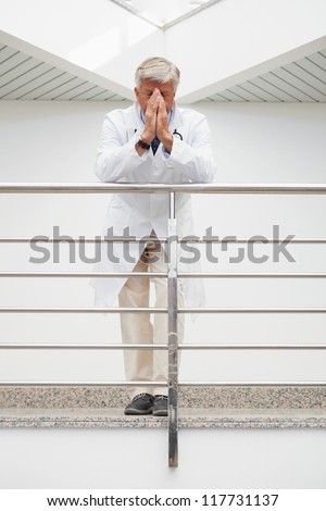 Worried doctor with face in hands leans against rail in hospital corridor