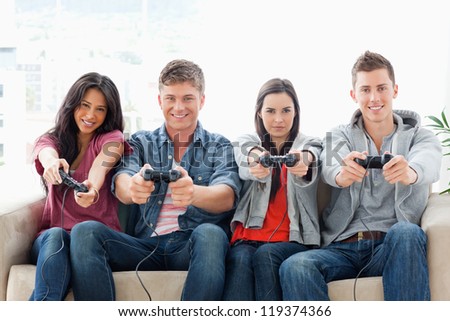 A smiling group of friends sit on the couch together while playing a game with controllers