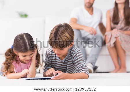 Brother and sister using tablet pc together on floor with parents behind them
