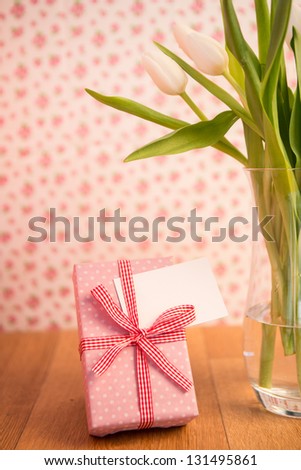 Vase of tulips on wooden table with pink wrapped gift and blank card on wallpaper background