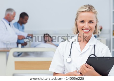 Nurse holding clipboard and looking at the camera in front of her team and a patient