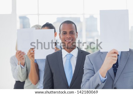 Businessman smiling at camera with colleagues covering faces with white pages