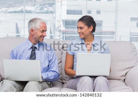 Business people using their laptops and smiling at each other on sofa in staffroom