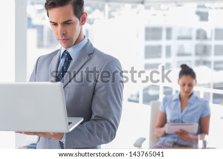 Businessman standing with laptop with woman working behind him