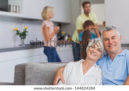 Elderly couple sitting on the couch and smiling at camera while their family is standing in the kitchen behind