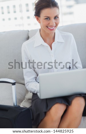 Businesswoman smiling at camera with laptop on the couch with suitcase