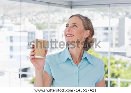 Happy businesswoman holding disposable coffee cup and looking up in bright office