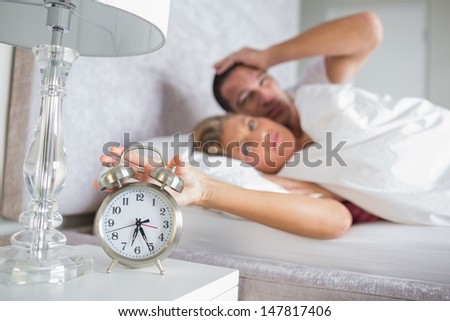 Tired couple looking at alarm clock in the morning with woman turning it off at home in bedroom