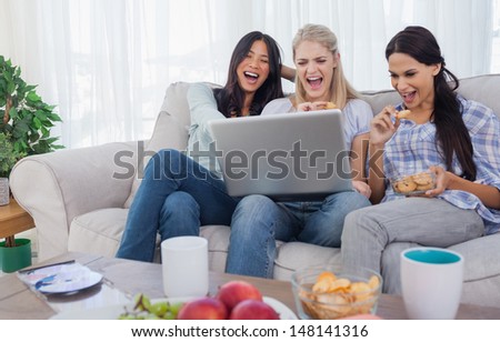 Smiling friends looking at laptop together and eating cookies at home on couch