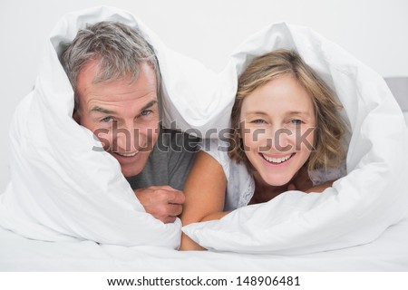 Happy middle aged couple under the duvet smiling at camera in bedroom at home