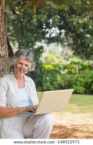 Happy mature woman using a laptop sitting on tree trunk smiling at camera in park