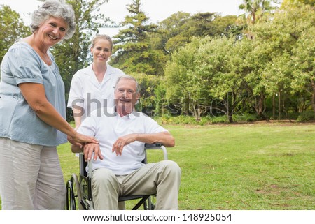 Cheerful man in a wheelchair with his nurse and wife smiling at camera in the park