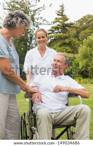 Happy man in a wheelchair talking with his nurse and wife in the park on sunny day