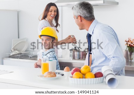 Boy trying on fathers hard hat with parents laughing at home in kitchen before work