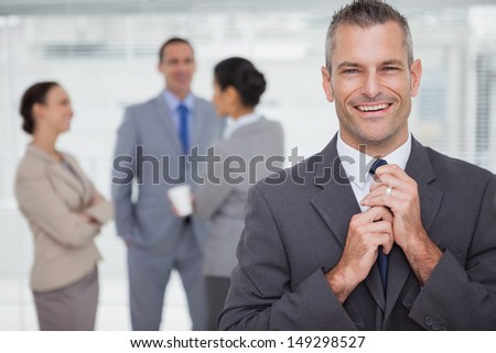Smiling manager tidying his tie up with employees in background in bright office