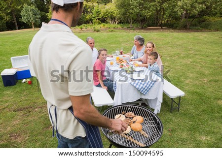 Father in chefs hat and apron cooking barbecue for his family sitting at picnic table