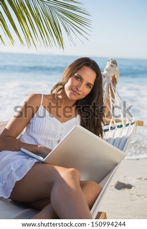 Brunette sitting on hammock with laptop smiling at camera on the beach