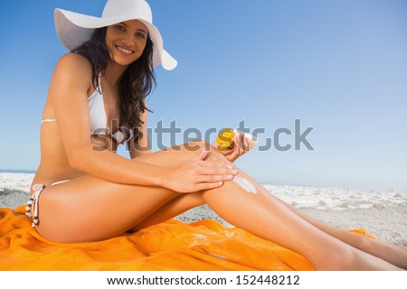 Cheerful young brunette with straw hat putting on sun cream on the beach