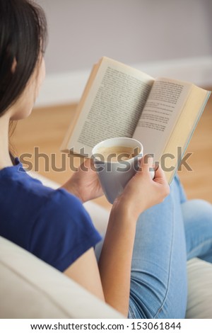 Young woman sitting on the sofa reading a book holding her coffee in living room at home