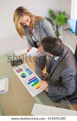 A smiling blonde businesswoman taking notes and explaining figures to a businessman sitting at his desk