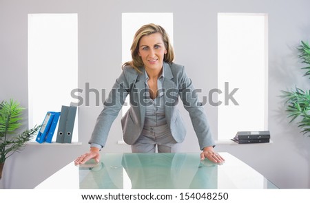Pleased blonde businesswoman standing firmly in front of a desk at office