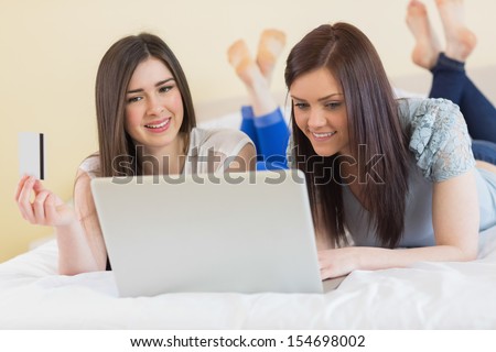 Cheerful friends using a laptop to shop online lying on bed at home in bedroom