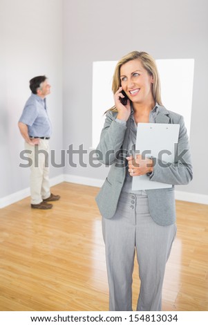 Smiling realtor calling someone with her mobile phone with buyer looking around the empty room