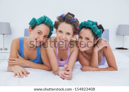 Girls in hair rollers lying in bed smiling and looking at camera at sleepover