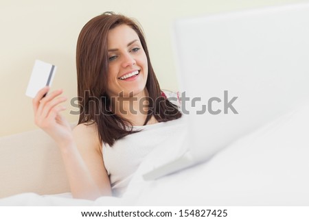 Excited girl using her laptop to shop online lying on her bed in a bedroom on a yellow background