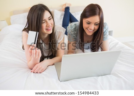 Smiling friends using a laptop to shop online lying on bed at home in bedroom