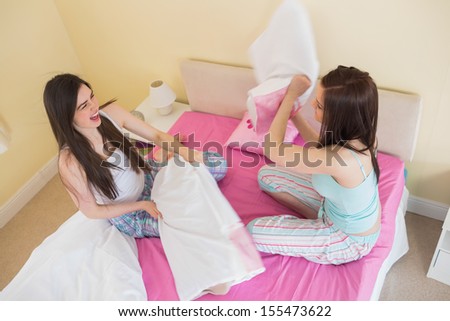 Happy friends in pajamas having a pillow fight on bed in bedroom at home