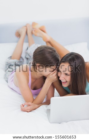 Friends lying in bed in pajamas with laptop and telling secrets