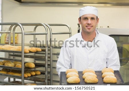 Happy young baker holding some rolls on a baking tray smiling at the camera