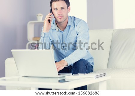 Content casual man using laptop and phoning in bright living room