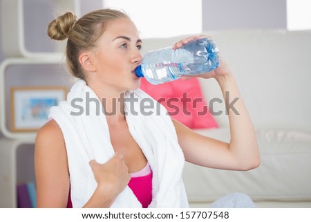 Thirsty blonde sitting on exercise mat drinking from water bottle in bright living room