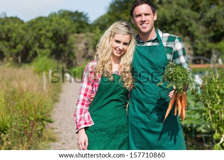 Cute couple posing in their garden holding carrots wearing aprons smiling at camera