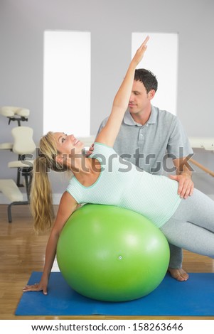 Smiling physiotherapist correcting patient doing exercise on exercise ball in bright room