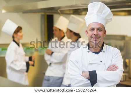 Mature male chef posing proudly in a kitchen with crossed arms and cooks in the background