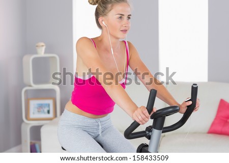 Sporty determined blonde training on exercise bike listening to music in bright living room