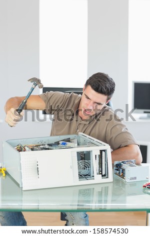 Handsome angry computer engineer destroying computer with hammer in bright office